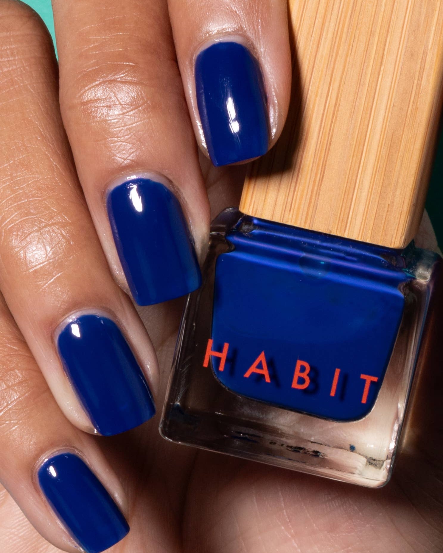 Hand with blue nail polish holding a bottle of 'Habit' nail polish. Night Drive Dark Blue Toxin Free Nail Polish