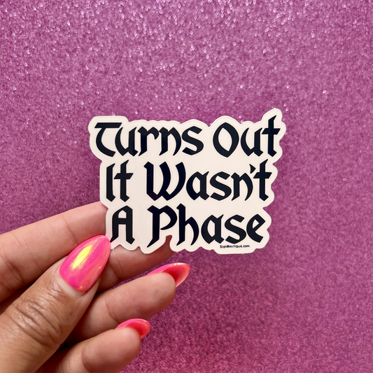 Hand holding a sticker with 'Turns Out It Wasn't A Phase' text against a pink glittery background