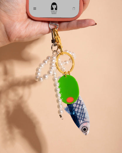 Keychain with green round charm and blue fish design held by a phone on a beige background