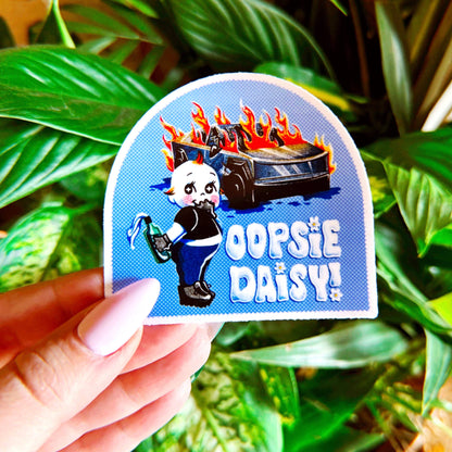 Sticker with cartoon character and text 'Oopsie Daisy' held by a hand against a leafy background. 