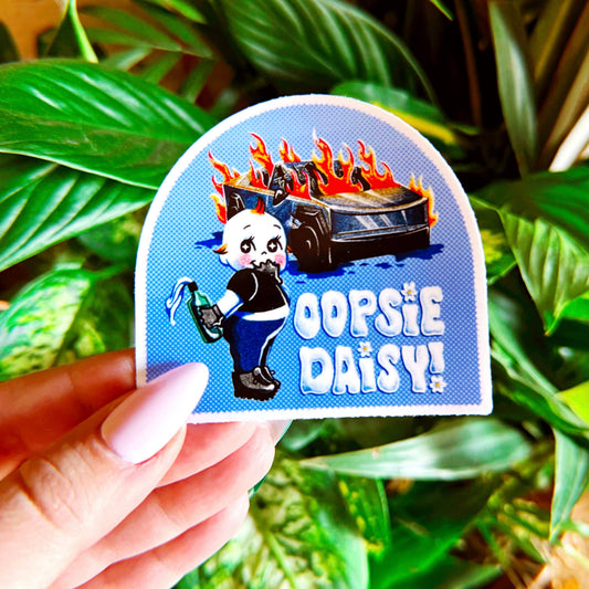 Sticker with cartoon character and text 'Oopsie Daisy' held by a hand against a leafy background. 