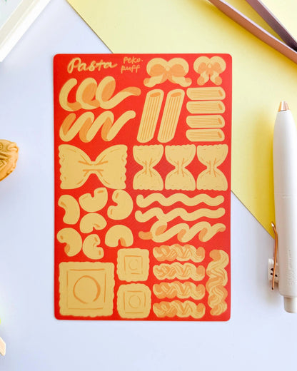 Pasta-themed sticker sheet on a red background with a pen and small toy on a light surface.