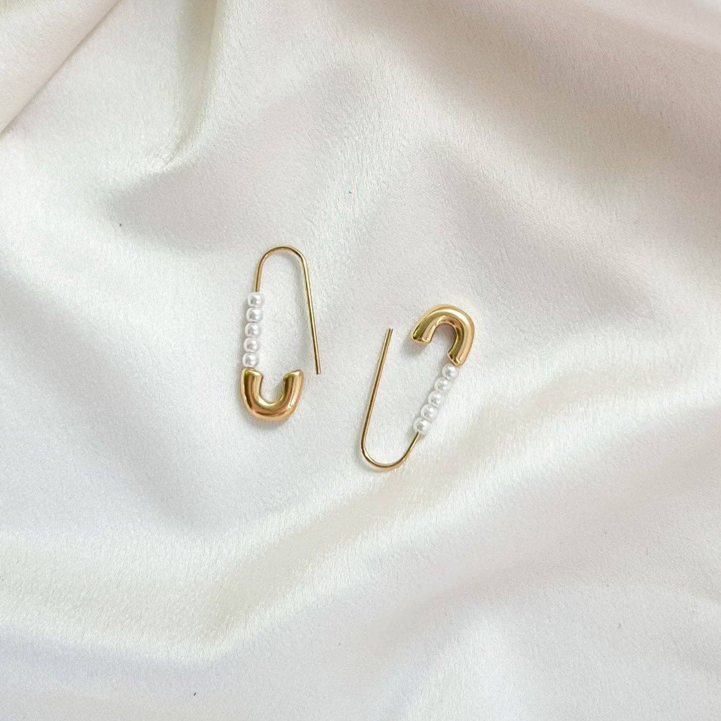 Gold safety pin earrings with pearl details on a white fabric background