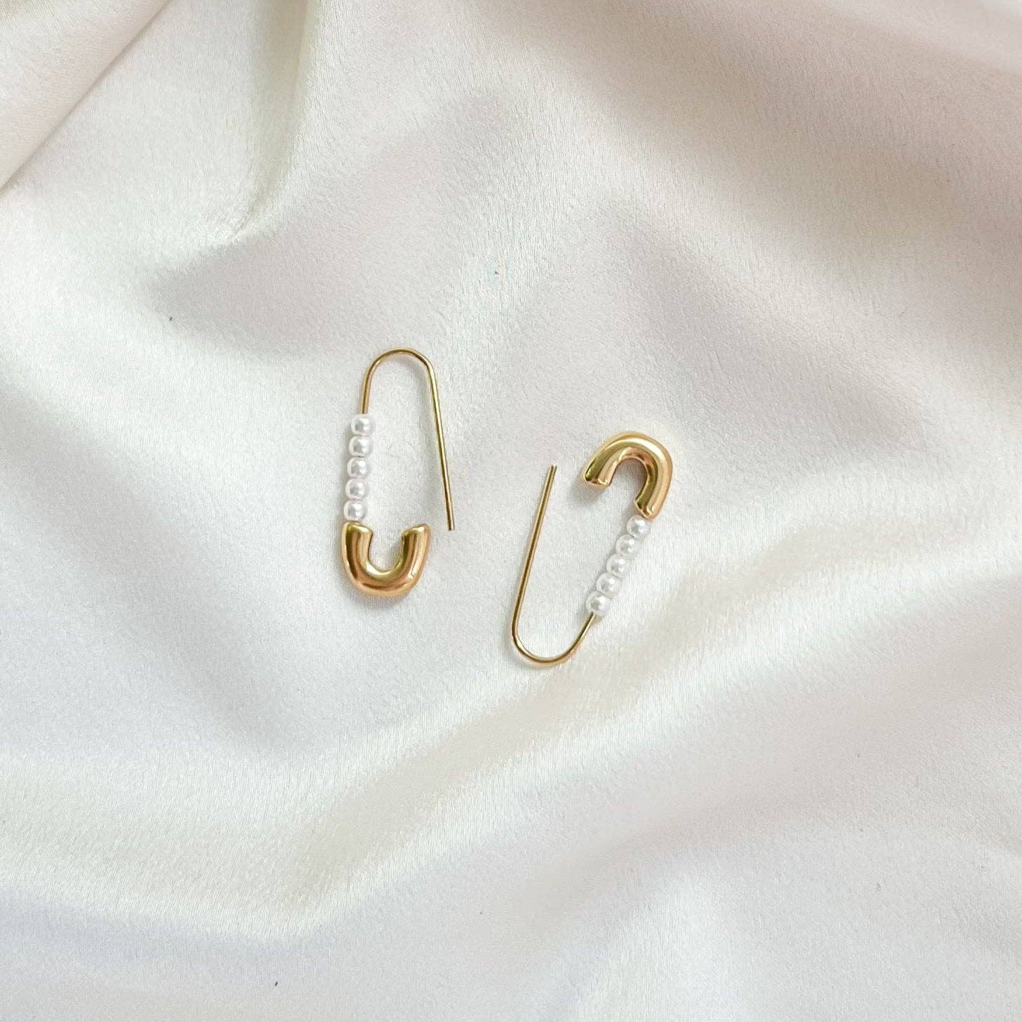 Gold safety pin earrings with pearl details on a white fabric background