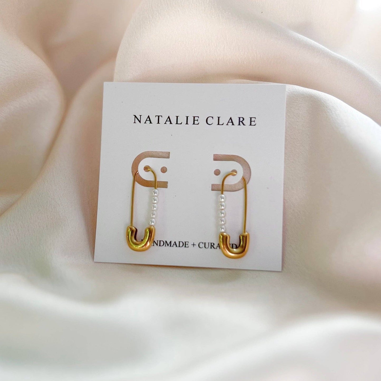 Gold safety pin earrings on a 'Natalie Clare' branded card against a beige fabric background