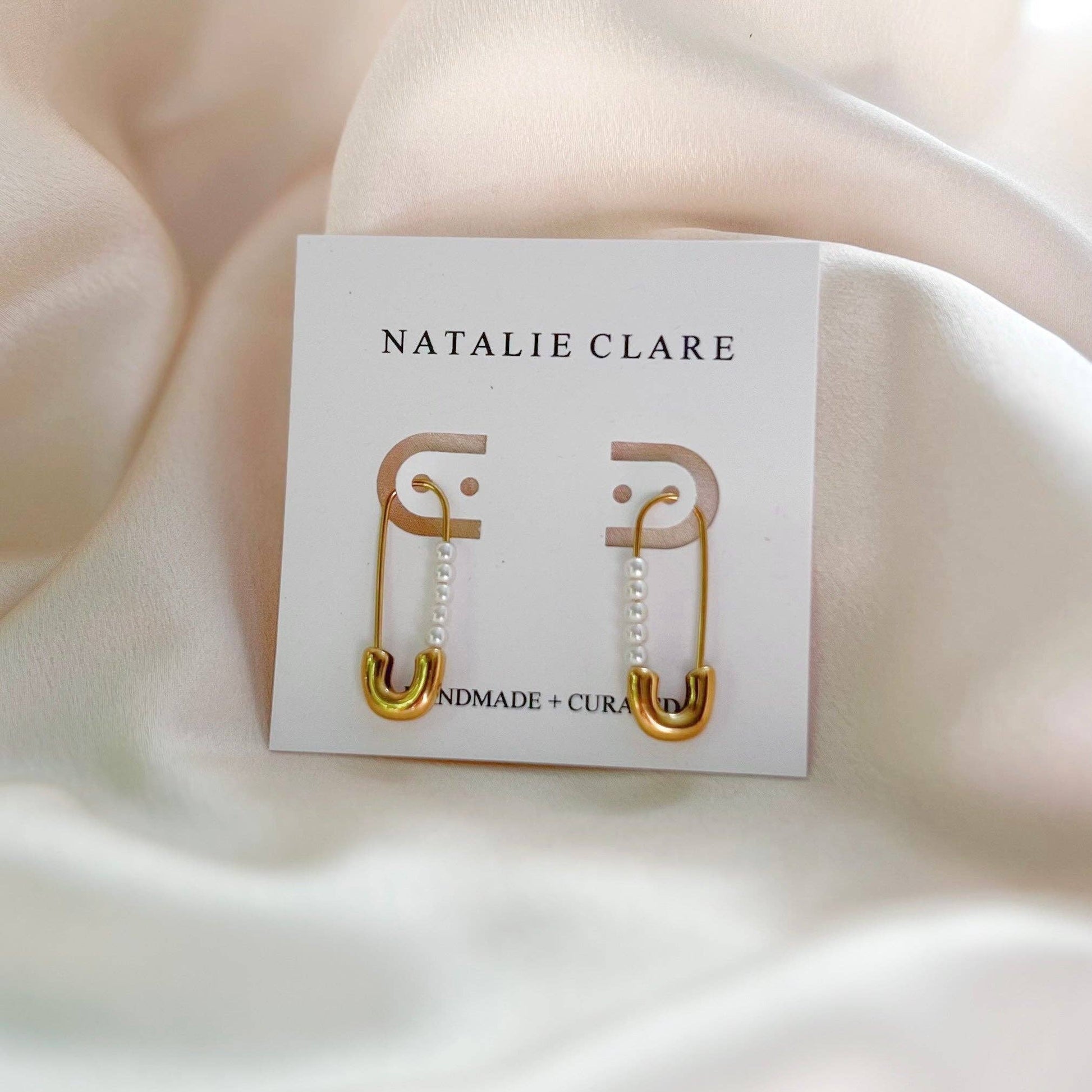 Gold safety pin earrings on a 'Natalie Clare' branded card against a beige fabric background