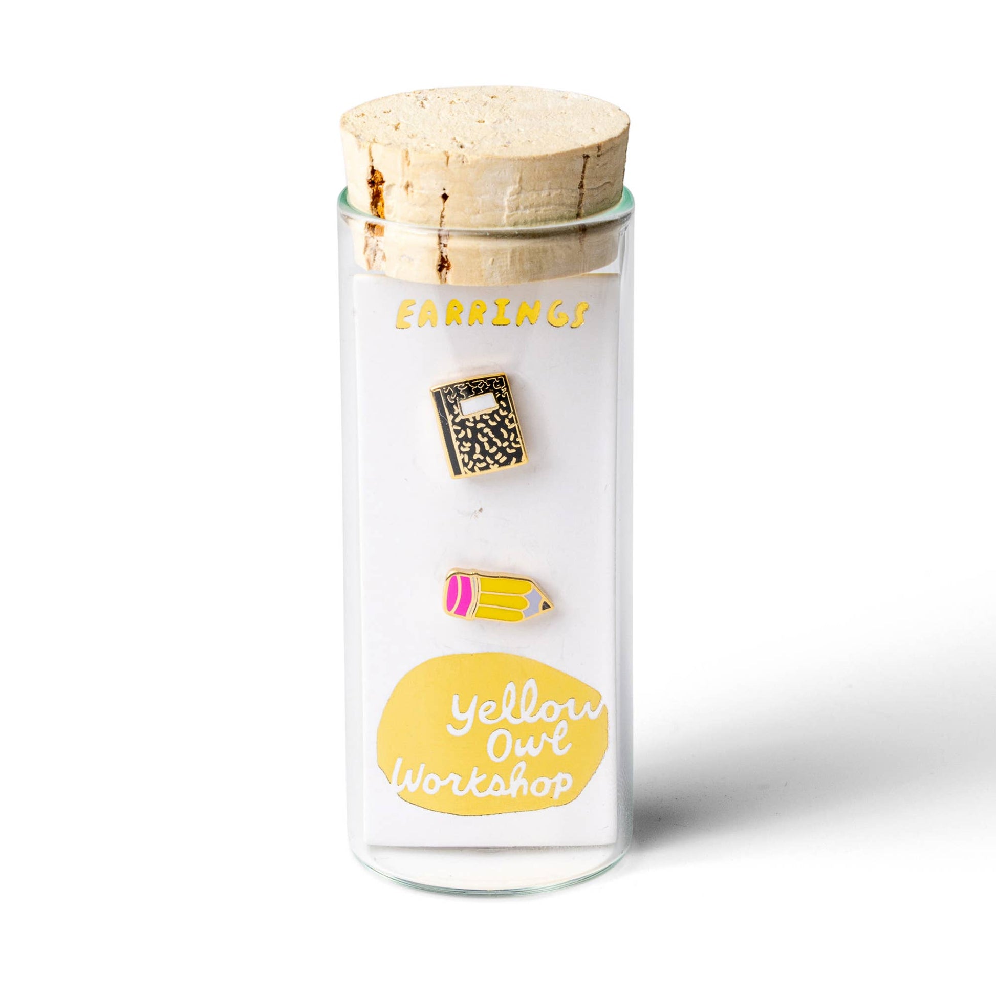 A pair of enamel pins shaped like a notebook and a pencil, displayed on a white card inside a clear glass jar with a cork lid. The card features the text 'EARRINGS' and 'Yellow Owl Workshop'.