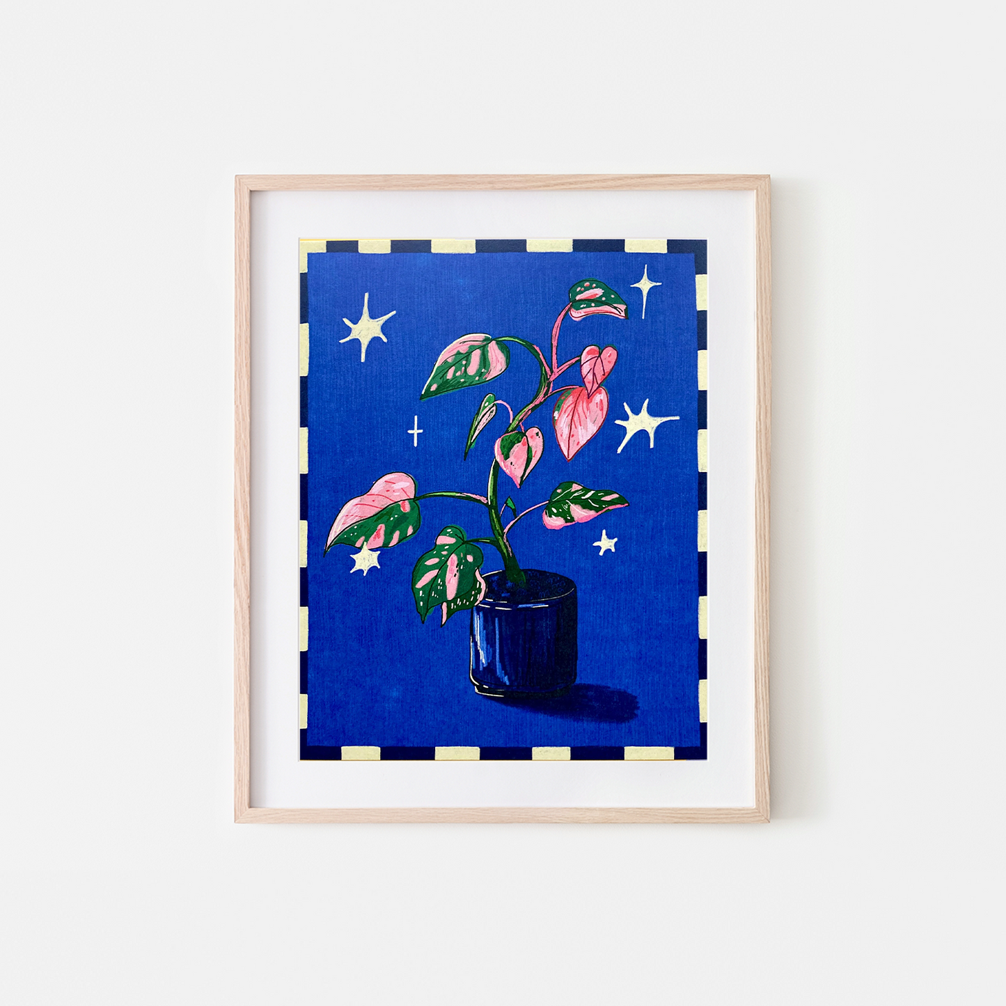 A framed art print featuring a potted plant with pink and green variegated leaves against a deep blue starry background, set in a light wood frame on a white wall.
