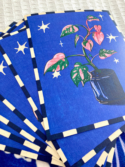 A stack of blue rectangular cards or art prints fanned out on a textured light surface. The top card shows a stylized plant with pink and green leaves in a dark pot, surrounded by white stars, while the others show matching blue backgrounds with navy-and-white striped borders.