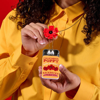 Person holding a red poppy necklace with a packaging container labeled 'California Poppy' by Mackbecks against a red background.