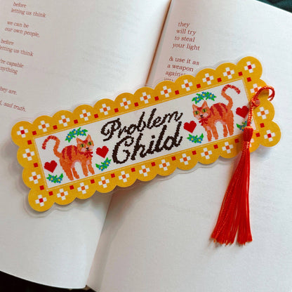 A decorative embroidered-style bookmark with a yellow scalloped border, two orange cats, red hearts, green leaves, and the words “Problem Child,” placed across open book pages with a red tassel hanging from the right side.