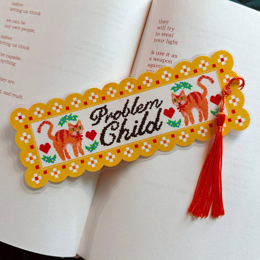 A decorative embroidered-style bookmark with a yellow scalloped border, two orange cats, red hearts, green leaves, and the words “Problem Child,” placed across open book pages with a red tassel hanging from the right side.