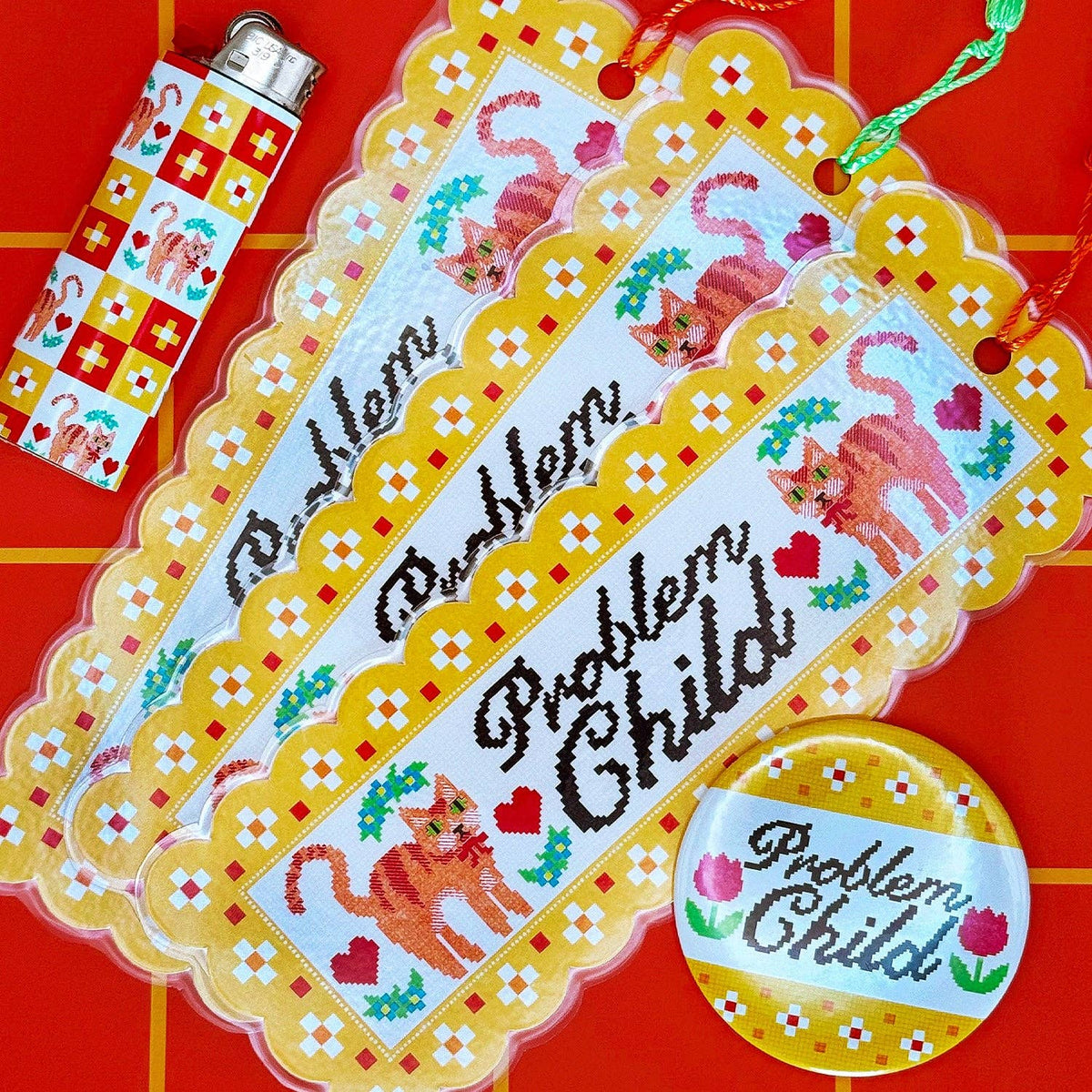 A set of novelty items printed with the phrase 'Problem Child' in script, including a large scalloped tag, a lighter, a round pin, and a small ribbon, all decorated with orange cats, red hearts, and white flowers on a bright red background.