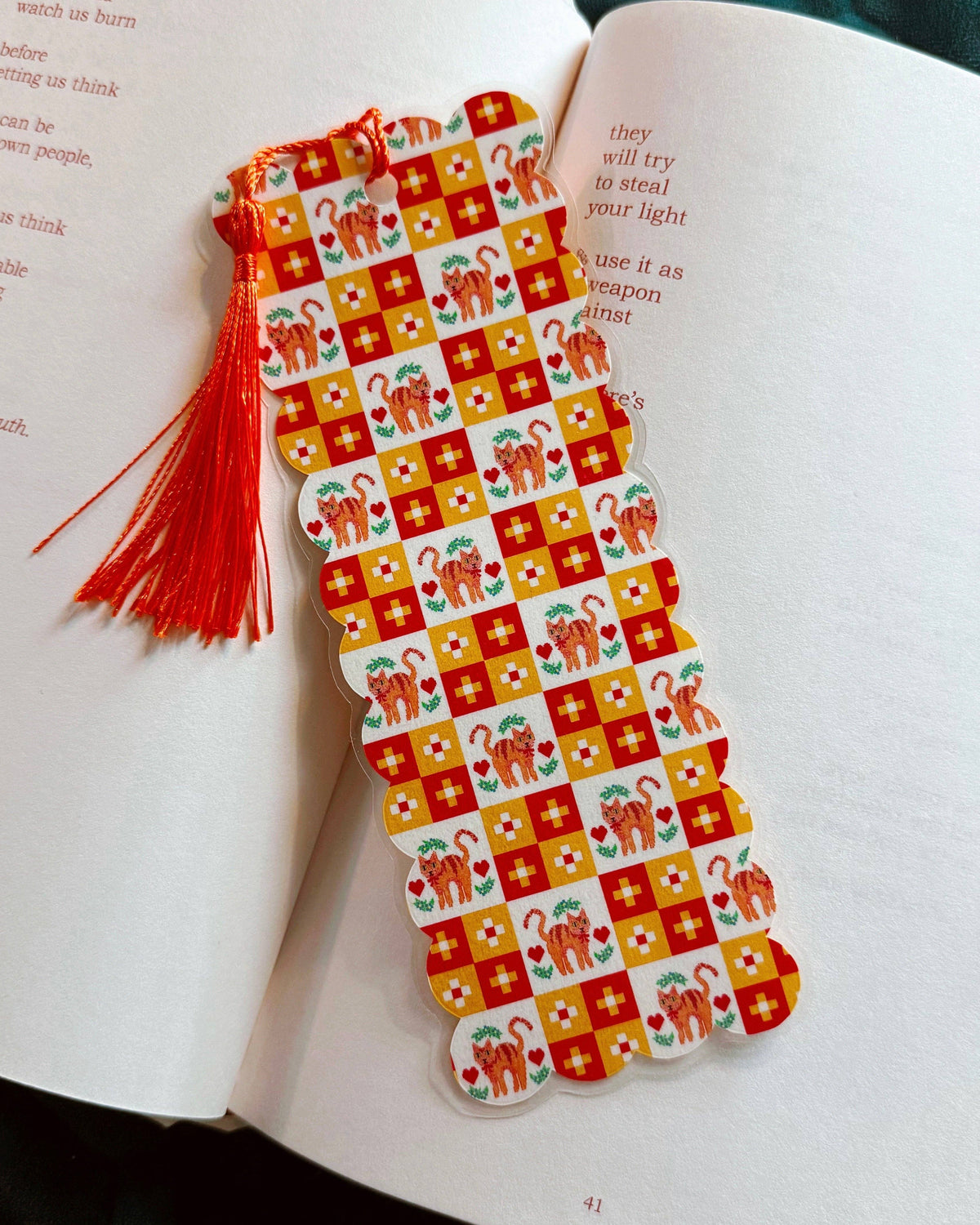 A decorative paper bookmark with a red and orange pattern of cats and flowers, featuring a red tassel, placed on the pages of an open book.