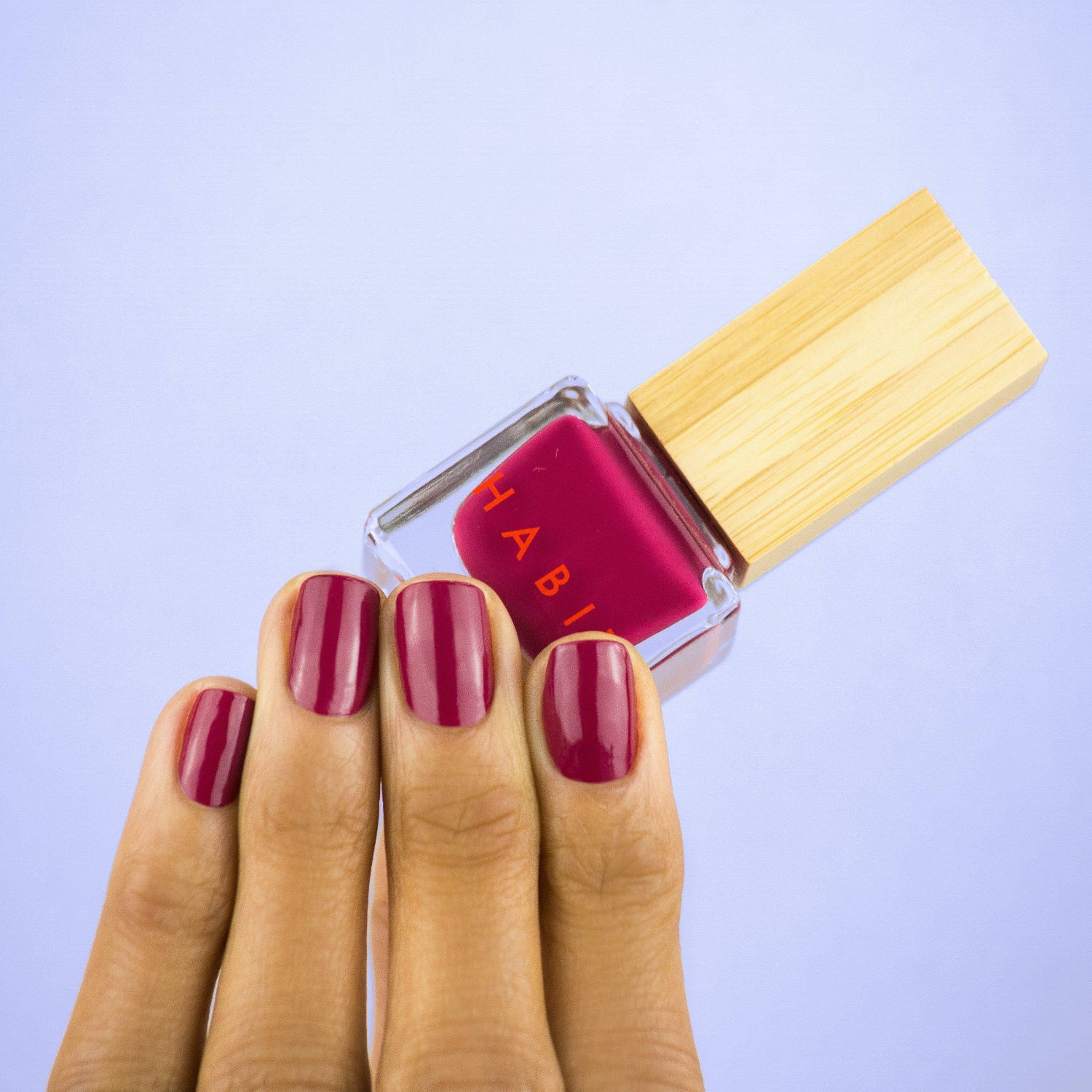 Nail polish bottle with wooden cap and matching red nails on a light purple background, R-Rated Red Toxin Free Nail Polish