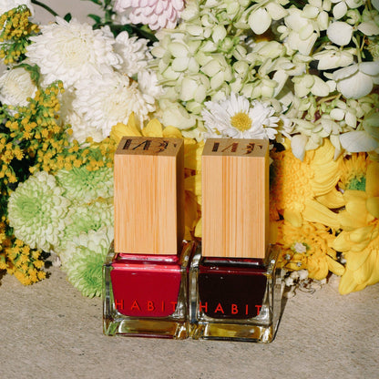 Two nail polish bottles with wooden caps and red and black polish, surrounded by flowers. R-Rated Red Toxin Free Nail Polish