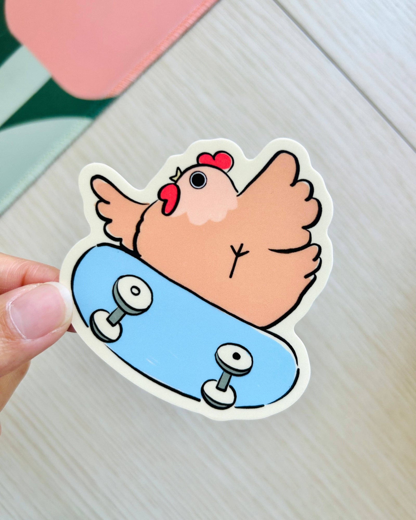 Cartoon chicken on a skateboard sticker held by a hand against a light background, Rad Skater Chicken Sticker