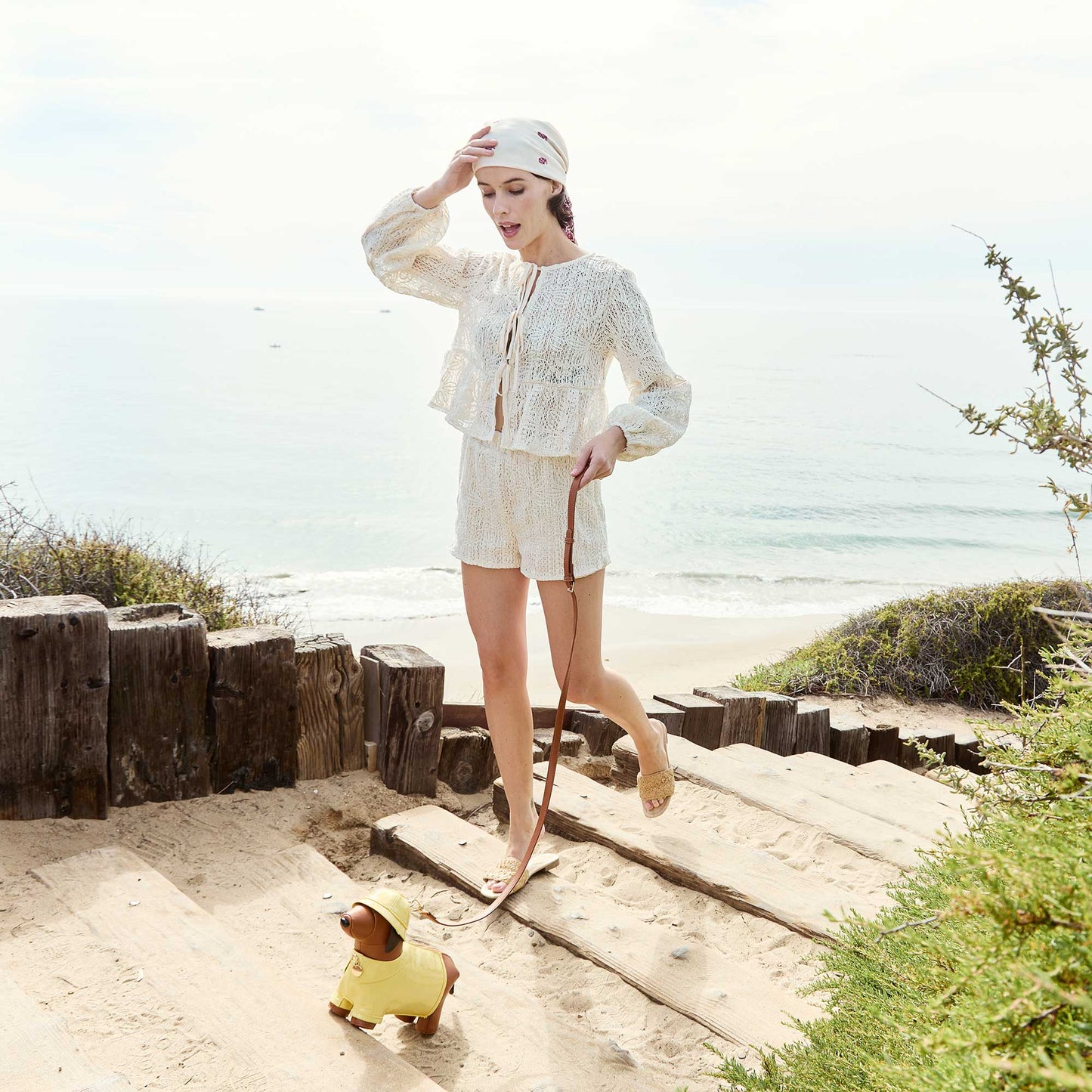 Woman in white outfit walking on a beach with a small dog in a yellow raincoat.