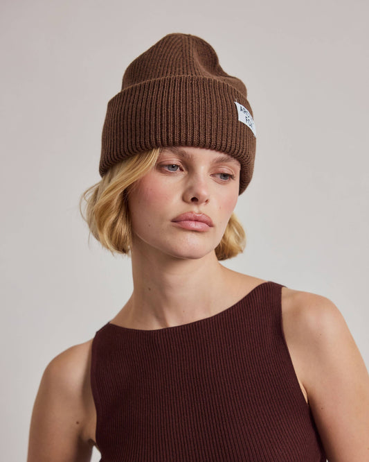 A woman wearing a brown ribbed knit beanie with a small white embroidered logo and a dark brown ribbed tank top, photographed against a plain light gray studio background.