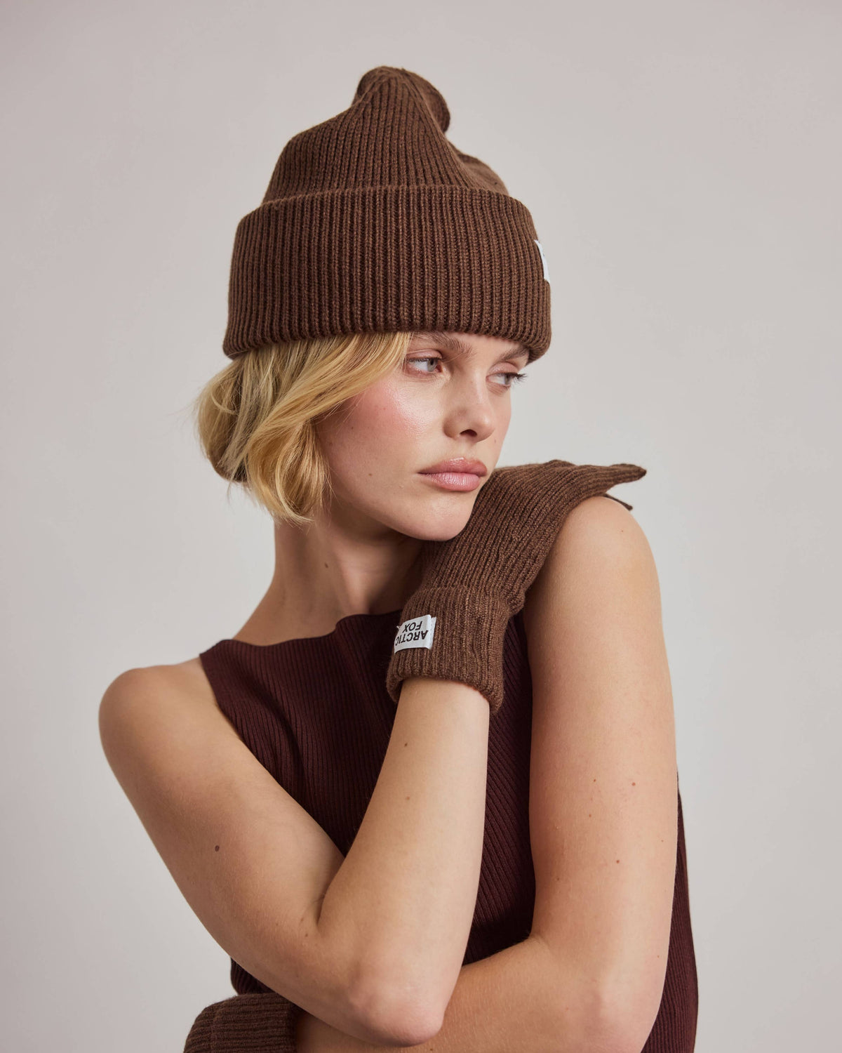 A woman wearing a brown ribbed knit beanie, matching brown knit mittens, and a dark brown ribbed knit top, posed against a neutral light gray studio background.