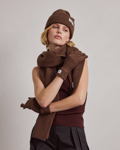 A model wearing a matching brown knit winter set including a beanie, scarf, gloves, and a sleeveless top, paired with black pants, against a plain light gray background.
