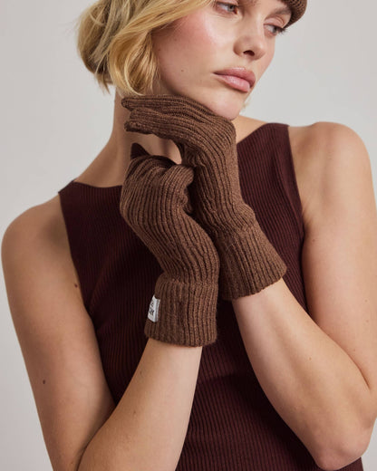 A woman wearing a brown ribbed knit beanie, matching brown knit fingerless gloves, and a dark brown ribbed tank top, posed against a light gray studio background.