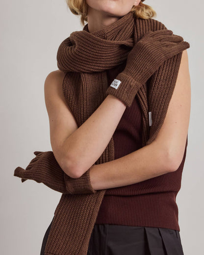 A model wearing a matching brown knit winter set including a beanie, scarf, gloves, and a sleeveless top, paired with black pants, against a plain light gray background.
