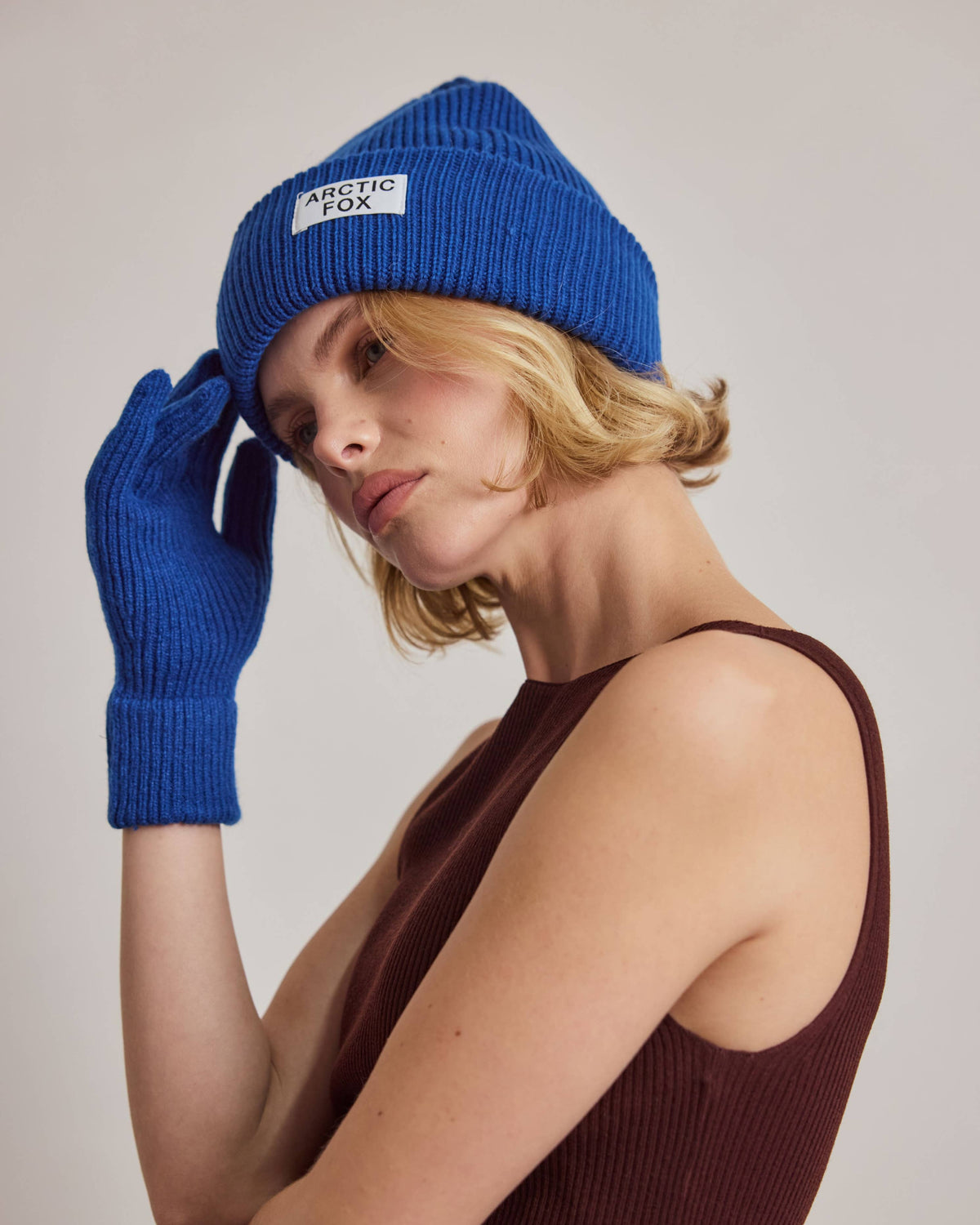 A woman wearing a bright blue knit beanie with a white 'METEOR FOX' label and matching blue knit gloves, posed against a neutral light gray background.