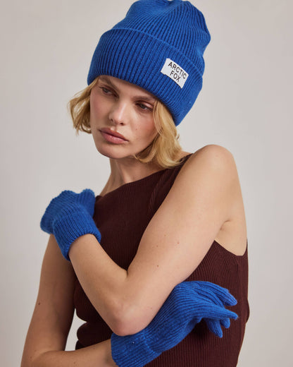 A model wearing a vibrant blue ribbed knit beanie with a white 'ARCTIC FOX' label, matching blue knit gloves, and a dark brown ribbed top, posed against a neutral light gray background.