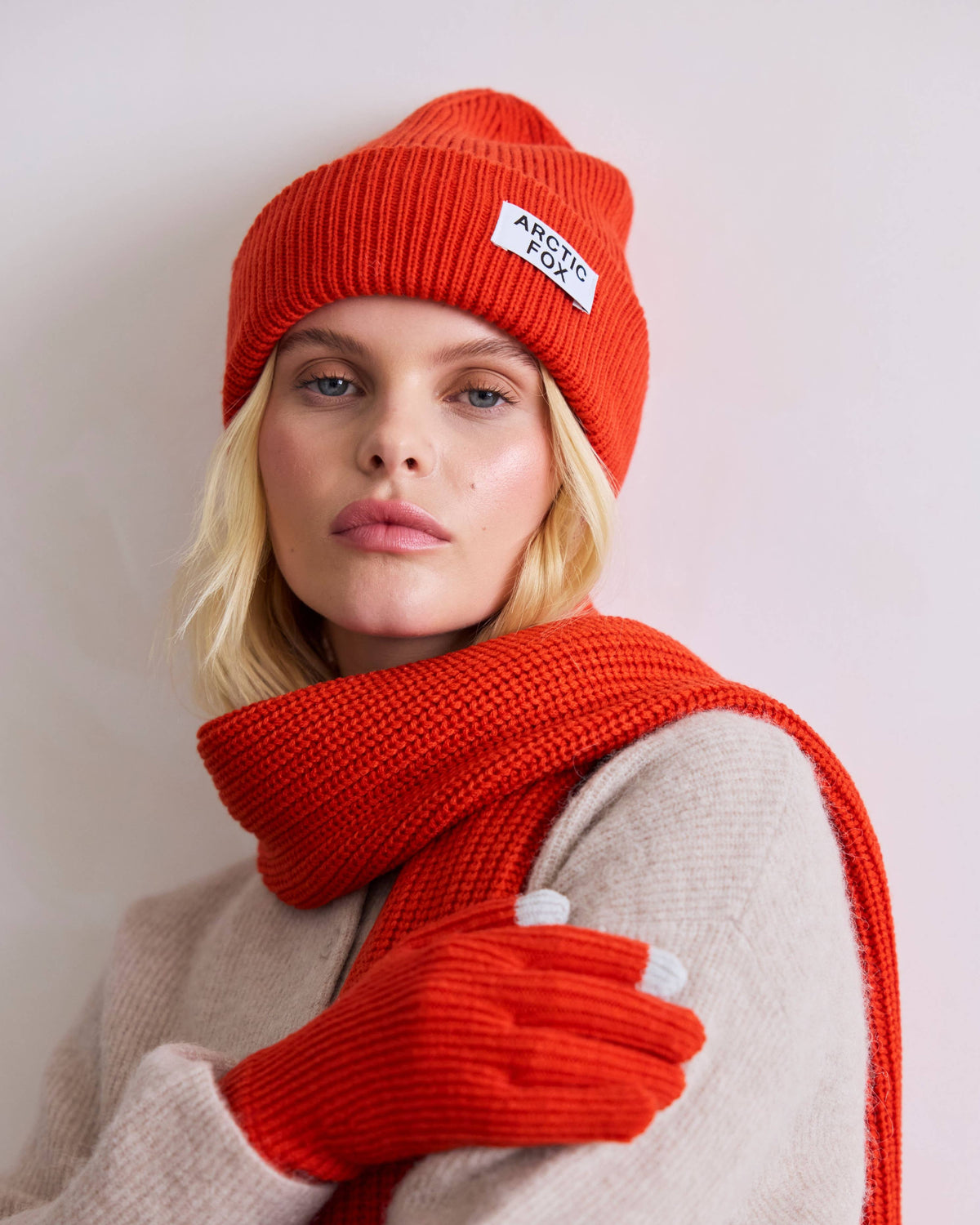 A woman wearing a bright orange-red knit beanie with a white 'Aberdeen' brand label, a matching knit scarf, and orange-red knit gloves, paired with a beige knit sweater against a plain light background.