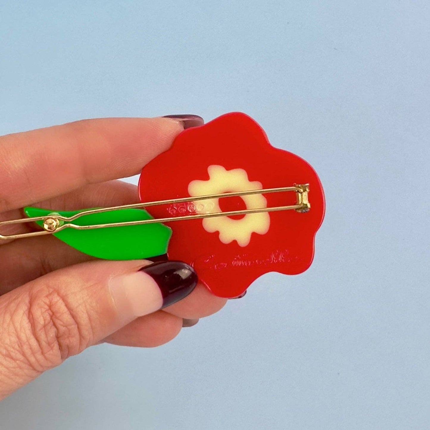 Red flower-shaped brooch with green leaves held by a hand against a light blue background, Red Flower Hair Clip