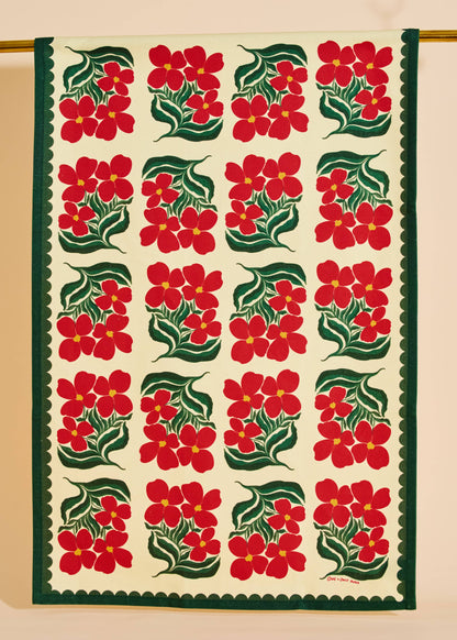 A square textile panel or tapestry with a repeating pattern of red flowers and green leaves on an off-white background, finished with a dark green scalloped border. The textile is hanging from a gold rod against a beige wall.