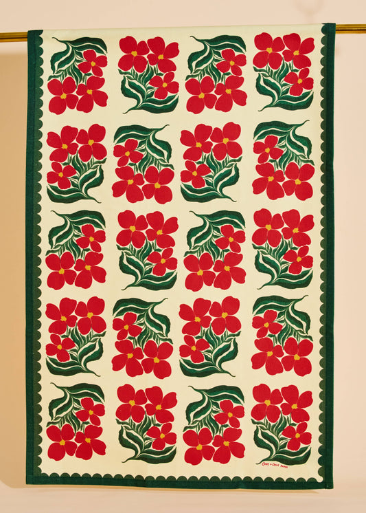 A square textile panel or tapestry with a repeating pattern of red flowers and green leaves on an off-white background, finished with a dark green scalloped border. The textile is hanging from a gold rod against a beige wall.