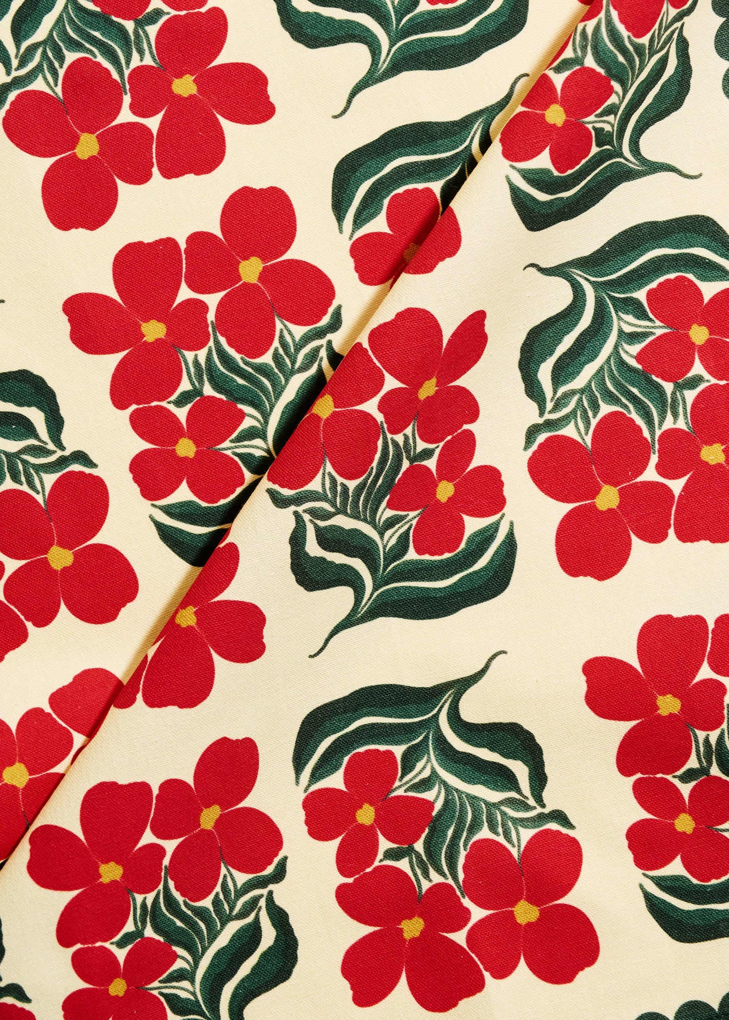A close-up of a fabric or wallpaper pattern featuring bright red five-petal flowers with yellow centers and dark green leaves on a warm cream background, divided diagonally by a thin gold-toned line.