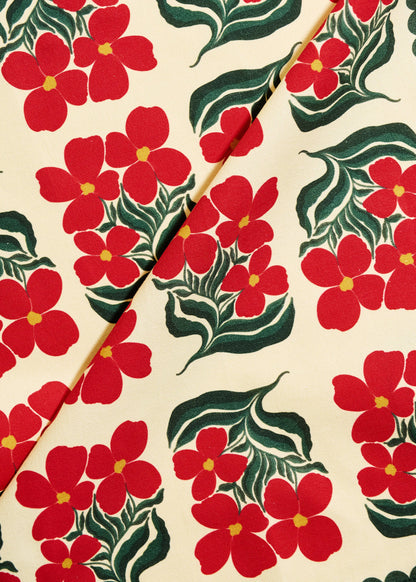 A close-up of a fabric or wallpaper pattern featuring bright red five-petal flowers with yellow centers and dark green leaves on a warm cream background, divided diagonally by a thin gold-toned line.