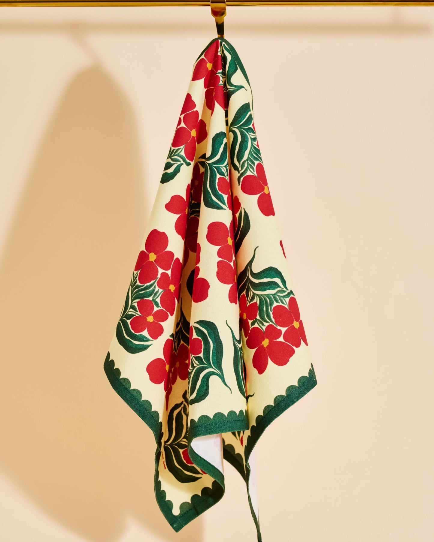 A cream-colored scarf with a bold red floral and green leaf pattern, hanging from a gold rod against a warm beige background.