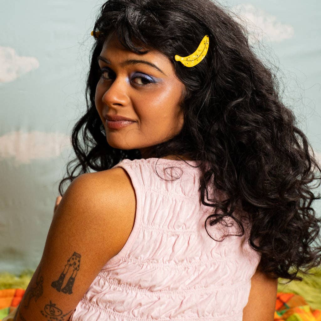 Woman with a yellow hair clip, pink sleeveless top, and tattoos on her arm against a blurred natural background.