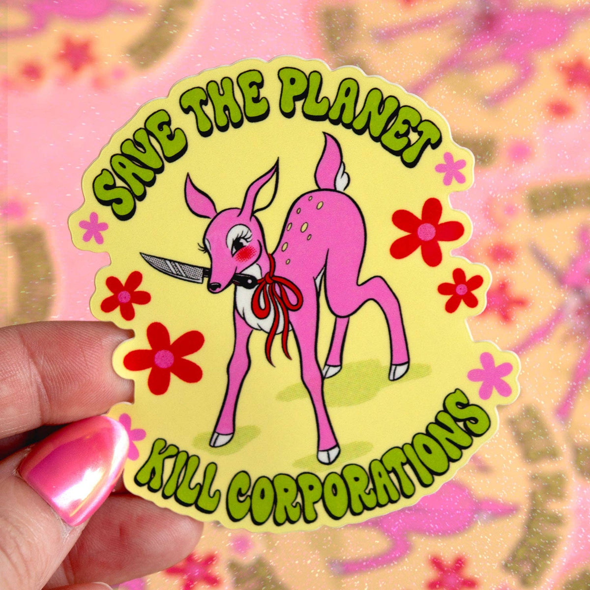 A hand with pink glittery nails holds a die-cut sticker featuring a pink cartoon deer holding a knife, surrounded by red and pink flowers, with the text 'SAVE THE PLANET KILL CORPORATIONS' in green bubble letters. The background is a blurry pink and gold patterned surface.