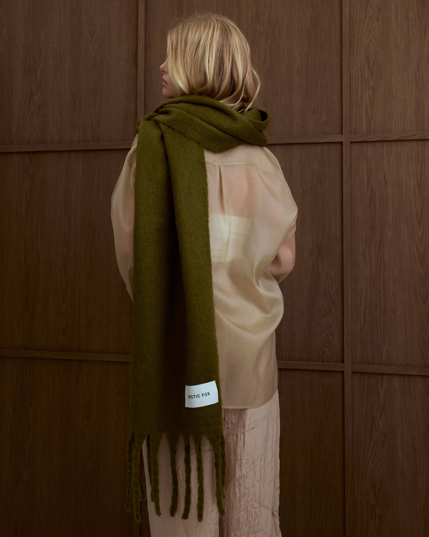 Person wearing a green scarf with a brand label against a wooden panel, Sea Kelp Green Reykjavik Recyled Bottle Scarf 