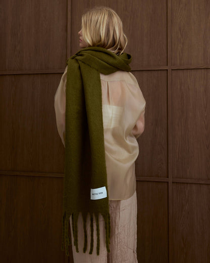 Person wearing a green scarf with a brand label against a wooden panel, Sea Kelp Green Reykjavik Recyled Bottle Scarf 