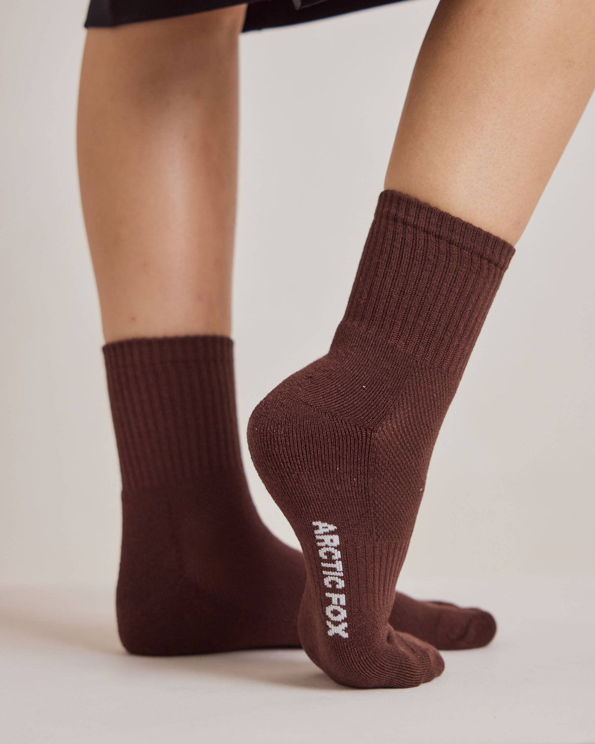 A close-up of a person's feet wearing dark brown ribbed crew socks with 'ARCTIC FOX' printed in white on the sole of one sock, set against a plain light beige background.