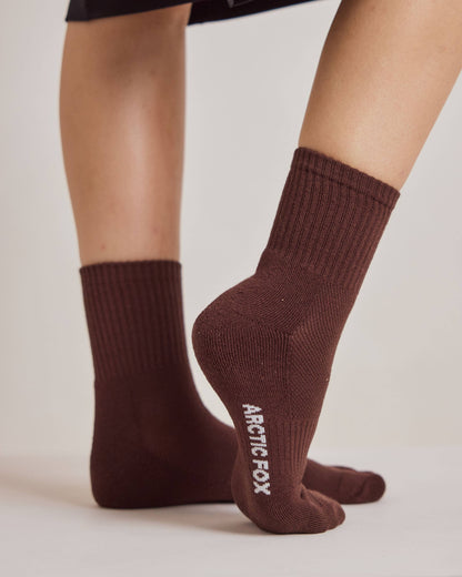 A close-up of a person's feet wearing dark brown ribbed crew socks with 'ARCTIC FOX' printed in white on the sole of one sock, set against a plain light beige background.