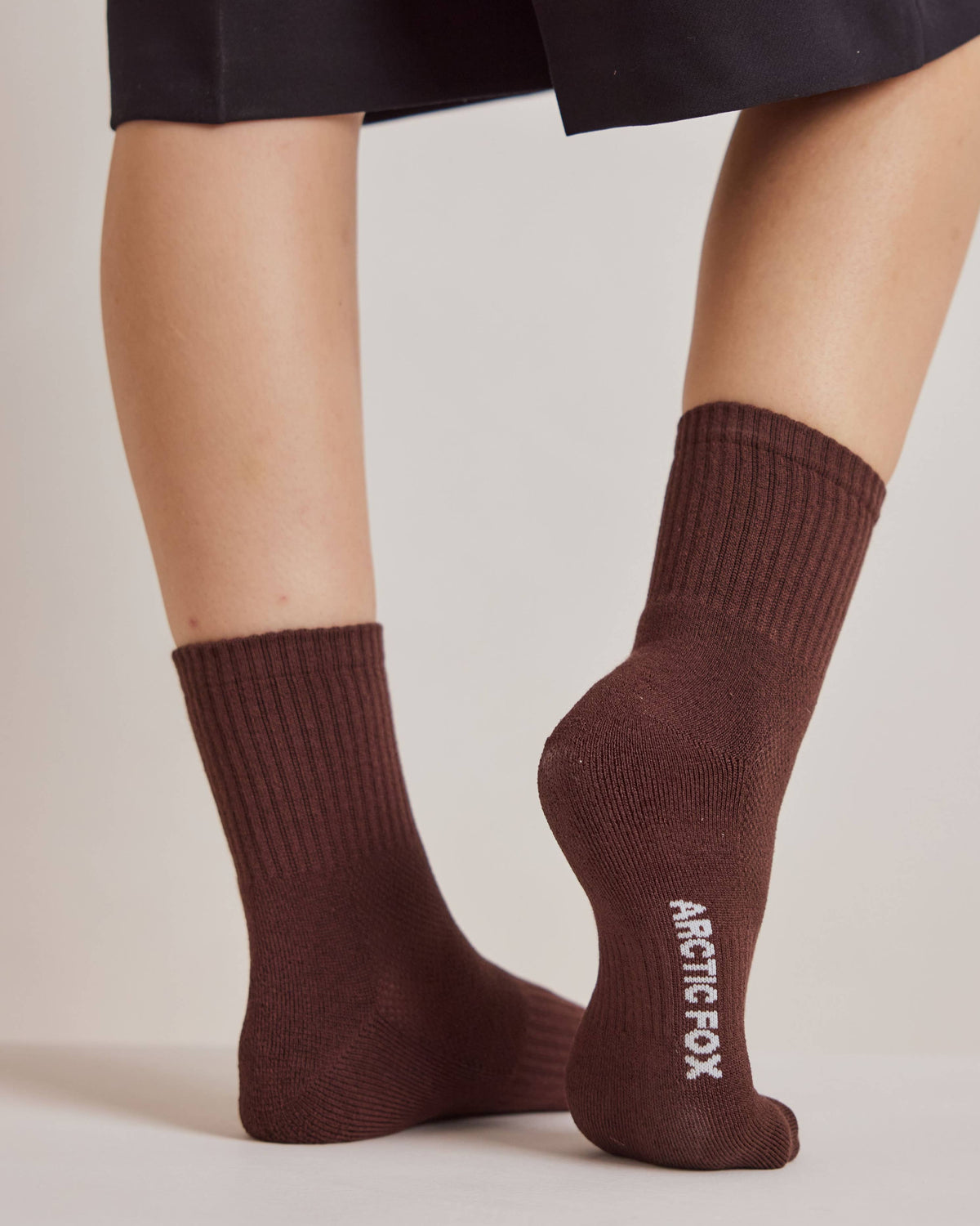 A close-up of a person's feet wearing dark brown ribbed crew socks with the brand name 'ARCTIC FOX' printed vertically in white on the sole of one sock, paired with black shorts against a light gray studio background.