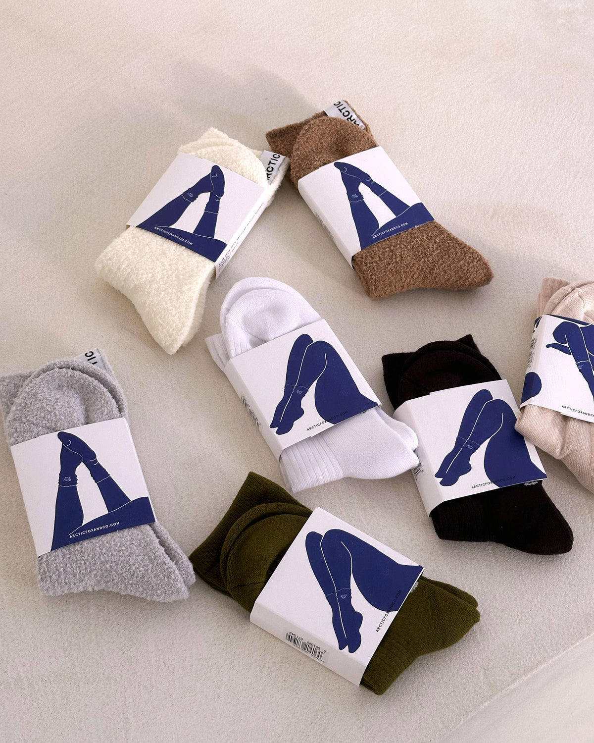 Six pairs of folded socks in different colors, each wrapped in a white paper band with a blue abstract graphic, arranged on a light beige fabric background.