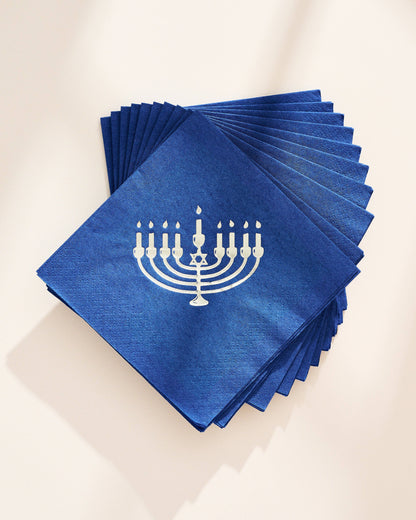 A stack of blue square paper napkins with a white printed menorah design, arranged in a fan shape on a light beige background.