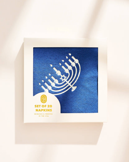 A square box of printed paper napkins on a warm off-white background with soft diagonal shadows. The napkin design shows a white menorah on a textured blue background, with a white lower-left label reading 'SET OF 20 NAPKINS' and smaller text below.