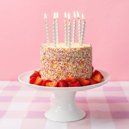 Birthday cake with colorful sprinkles and lit candles on a white cake stand against a pink background.