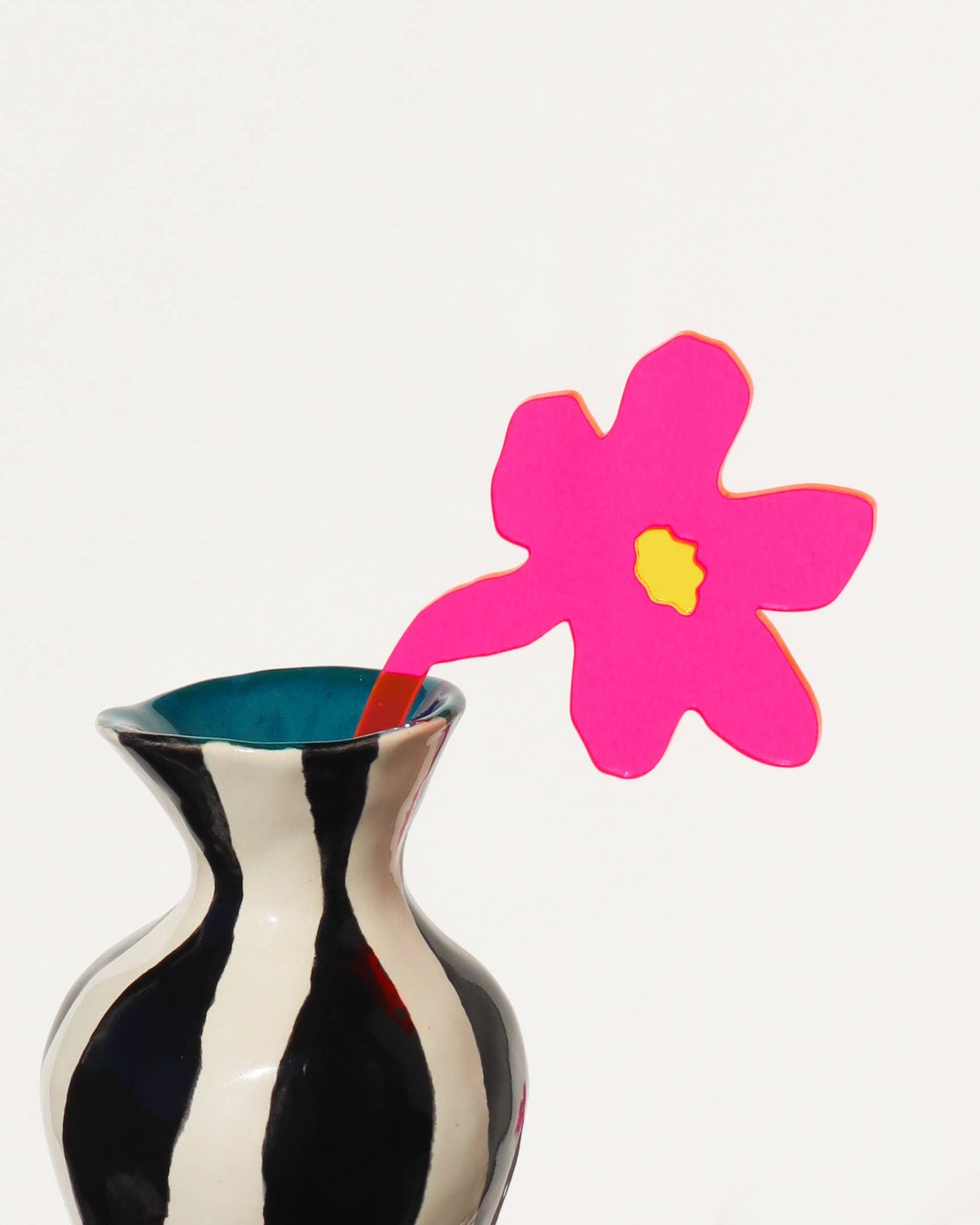 A bright pink paper flower with a yellow center inserted into a glossy black and white striped ceramic vase with a teal interior, set against a plain off-white background.