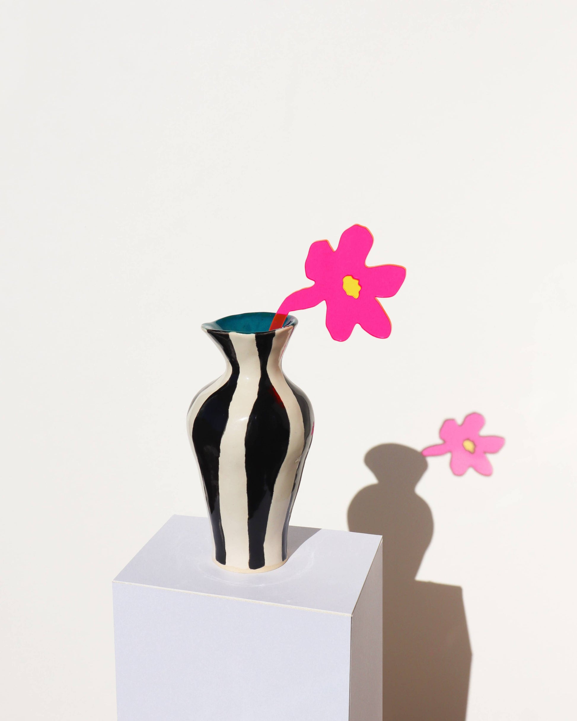 A black and white vertically striped ceramic vase with a bright blue interior, holding a single pink paper flower with a yellow center. The vase sits on a white pedestal against a white wall, casting a sharp shadow to the right.