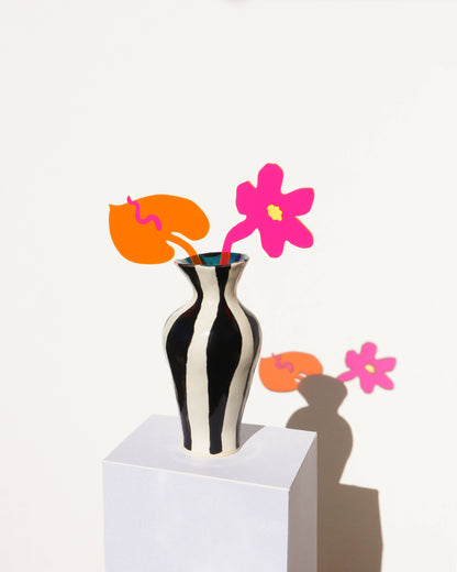 A black and white vertically striped ceramic vase holding two stylized paper flowers in pink and orange, displayed on a white pedestal against a white wall with a sharp shadow.